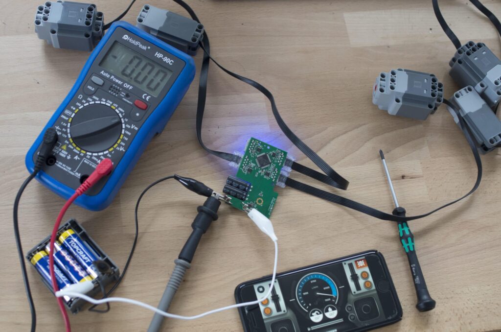 Messgeräte für physische Prototypen. Man sieht ein Messgerät, Batterien, eine mit Kabeln verbundene Platine einen Schraubendreher und ein Smartphone.