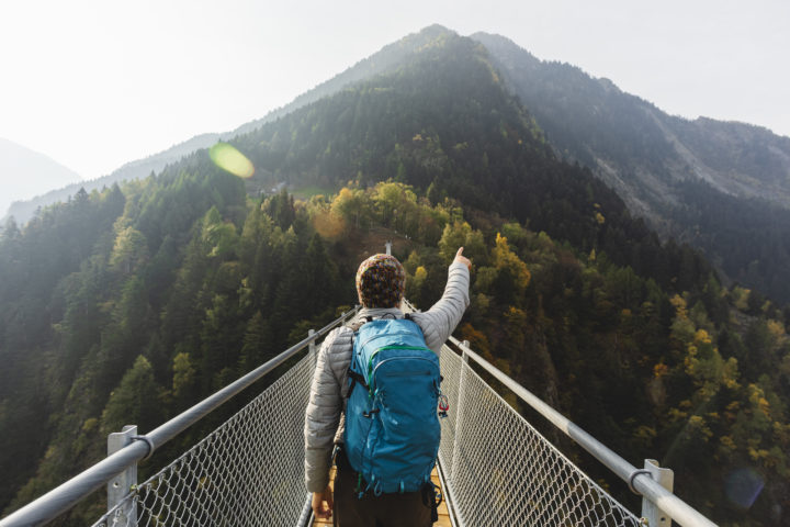 Person auf einer langen Brücke mit der man einen Berg erreichen kann. Die Person zeigt mit ihrer Hand auf die Berglandschaft