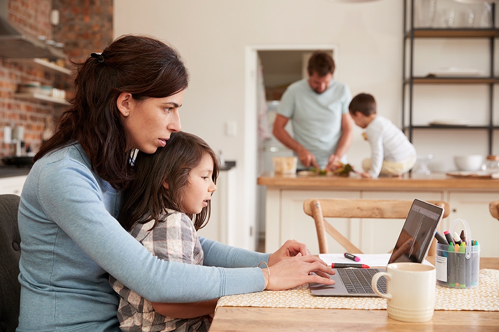 Eine Frau sitzt mit ihrem Kind am Laptop und beide schauen auf den Bildschirm. Im Hintergrund steht der Vater in der Küche und der Sohn schaut zu