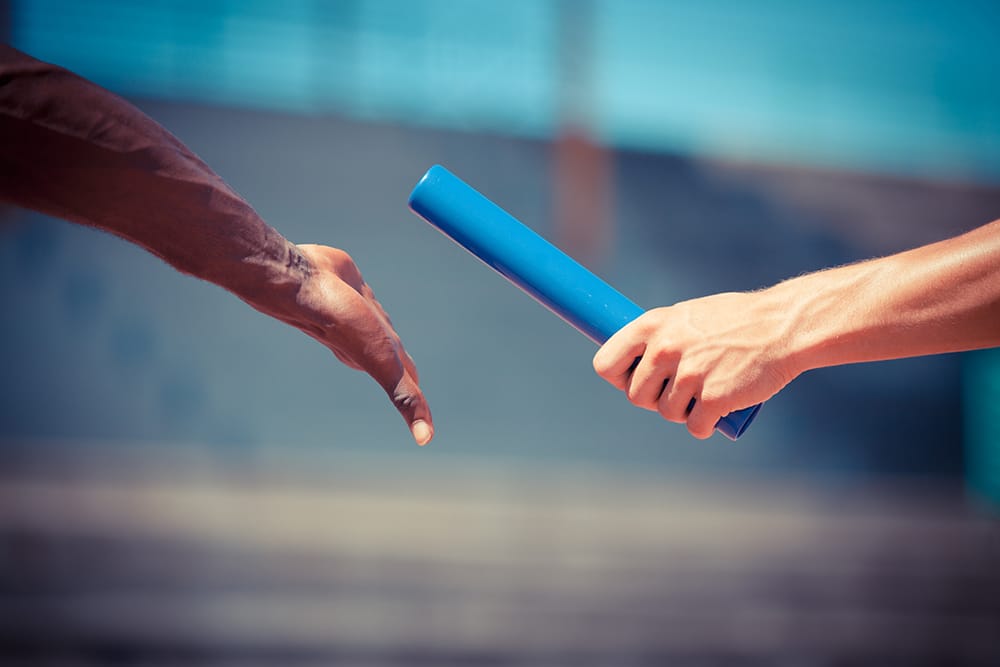 Man sieht zwei zwei Hände. Die eine Hand übergibt der anderen einen blauen Staffelstab